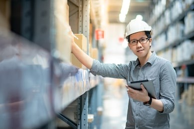 young asian man worker wearing safety helmet and eyeglasses doing stocktaking of product in cardboard box on shelves in warehouse by using digital tablet and pen. physical inventory count concept