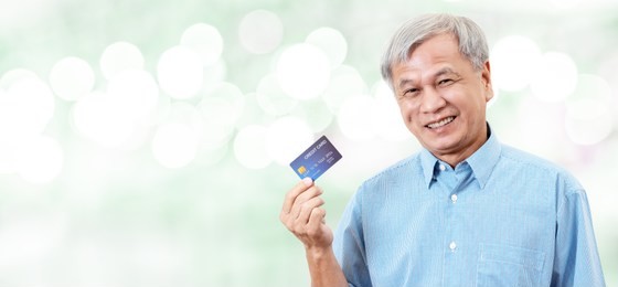portrait of happy senior asian man holding credit card and showing on hand smiling and looking at camera on isolated blurred bokeh background banner feeling positive or enjoy. older lifestyle concept.