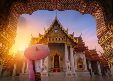 woman holding traditional red umbrella on the marble temple, wat benchamabopitr dusitvanaram at sunrise in bangkok, thailand.