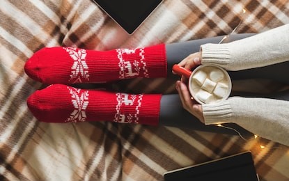 women's hands and feet in sweater and woolen cozy red socks holding cup of hot coffee with marshmallow, sitting on plaid with garland, tablet and notebook. concept winter comfort, morning drinking