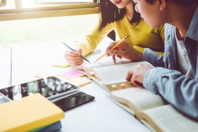 high school or college using laptop while sitting at table. group students studying and reading with books in library. students helps friend catching up and learning tutoring. education concepts
