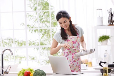 young woman in kitchen cooking with reference the laptop