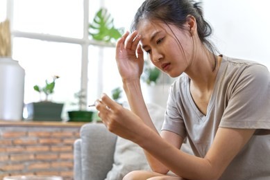 young asian women on sofa suffering from headache and have some fever. she is holding thermometer on left hand and right hand is touching her face. medical and healthcare concept.