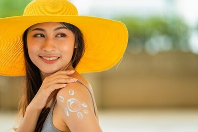 portrait of a beautiful smiling asian woman in yellow hat with sun shape created from sunscreen lotion on her shoulder outdoor. copy space
