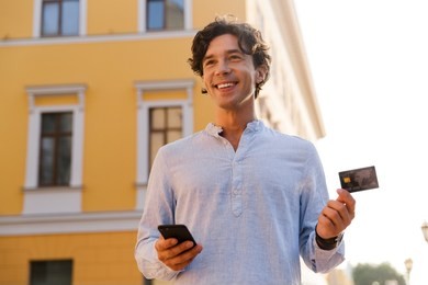 cheerful young casual man holding mobile phone while walking at the city street, holding credit card