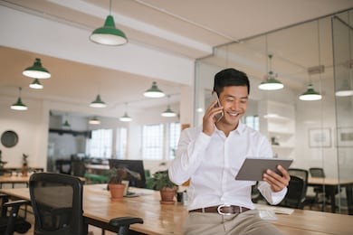 smiling young asian businessman sitting on a desk talking on a cellphone and using a digital tablet while working alone in a modern office