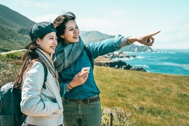 close friends looking into the ocean and one girl pointing to somewhere with a phone held by a background of stunning coast.