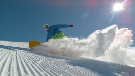 close up: extreme snowboarder carving downhill and spraying snow into camera. smiling active man snowboarding on groomed ski slope in mountain resort. cheerful snowboarder turning