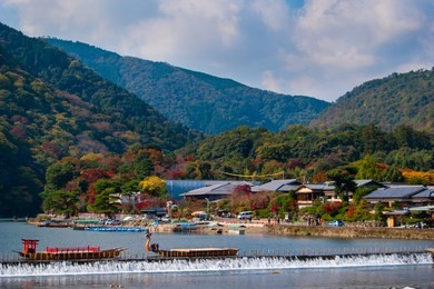 the view of  katsuragawa river, mountains covered with green, yellow, orange and red trees, and traditional boats. shot in arashiyama in kyoto during momiji season on a sunny day