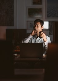 young asian businessman working at his desk in an office late at night yawning while drinking a cup of coffee to stay awake