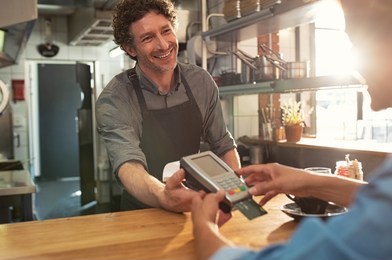 woman paying by credit card and entering pin code on reader holded by smiling barista in cafeteria. customer using credit card for payment. mature cashier accepting payment over nfc technology.