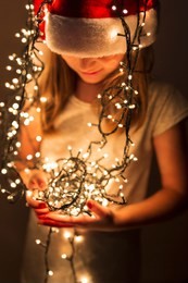 beautiful little girl wearing santa hat and holding bunch of christmas lights; child setting up christmas decorations and waiting for santa