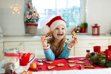 kids bake christmas cookies. child in santa hat cooking, decorating gingerbread man for xmas celebration. family preparing sweets in white kitchen with christmas tree on snowy winter day.
