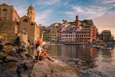 young woman sitting on a rock watching sunset at the harbour of vernazza cinque terre italy, sunset at harbor people watching village vernazza cinque terre