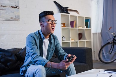 excited young asian man yelling and playing video game with joystick at home 
