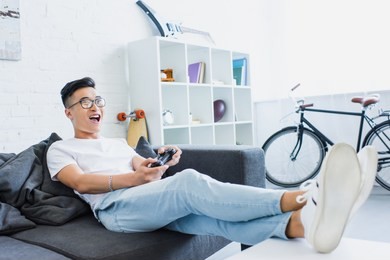 excited handsome asian man playing video game on sofa in living room