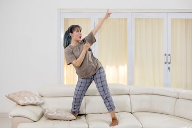 young woman singing with a remote control while dancing on the couch. shot at home
