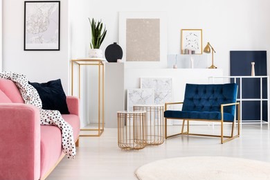 silver abstract painting on the wall of trendy living room with two elegant coffee tables, petrol blue armchair and powder pink couch with doted blanket on it