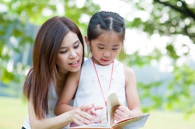 beautiful little girl reading book with her mother and smiling. summer park in background.