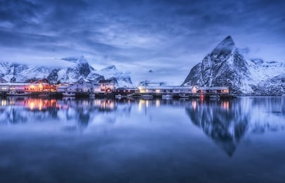 fishing village with boats at night, lofoten islands, norway. winter landscape with buildings, illumination, snowy mountains, sea, purple cloudy sky reflected in water at dusk. norwegian rorbuer