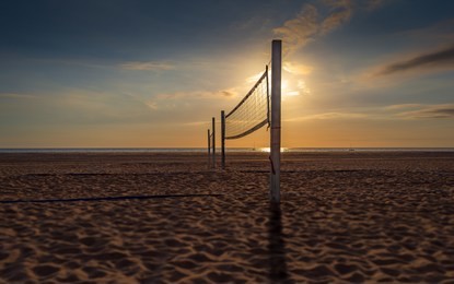 a beach volleyball court at sunset, the courts are in silhouette against the setting sun.  the sun, clouds, sky, boats and water are all visible