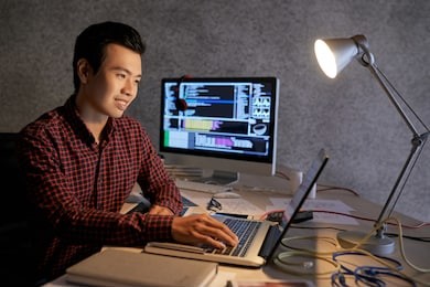 smiling young asian it student working on laptop at table with various cords and documents