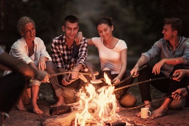 a group of happy young friends relaxing and enjoying  summer evening around campfire on the river bank