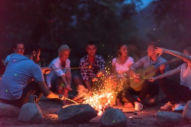 a group of happy young friends relaxing and enjoying  summer evening around campfire on the river bank