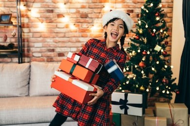 cute girl holding gift showing big toothy smile. lovely daughter with santa hat standing in front of the xmas tree. joyfully getting gifts at christmas holiday at home.
