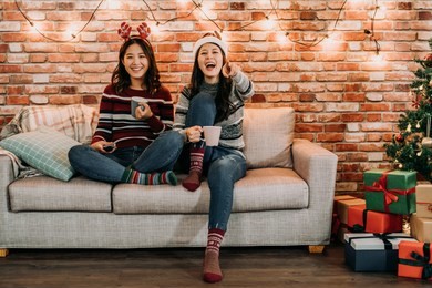 friends watching comedy at home on christmas eve. young girl laughing pointing at the television. comfortable sitting on sofa in xmas holidays lifestyle.