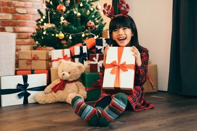 little girl with deer on head got a big present as christmas gift. child holding gift with red ribbon showing big toothy smile. lovely daughter being joyful, fresh and cheerful.