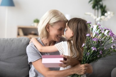 happy grandmother and granddaughter hug celebrating birthday together, cute little girl congratulate granny presenting gift box and flowers bouquet, grandma thanking grandchild for surprise
