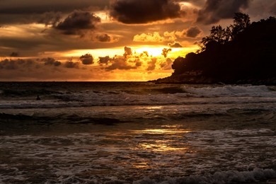 a spectacular orange sunset with dramatic dark clouds off the north point of karon beach, phuket thailand. a scene of paradise with tropical vegetation on the distant coast and calm  waters.