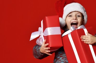 funny smiling joyful  child boy in santa red hat holding christmas gift in hand over the red background