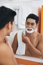 smiling young asian man shaving in front of bathroom mirror