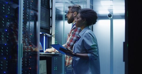 modern multiethnic man and woman with tablet using laptop in server room while checking servers