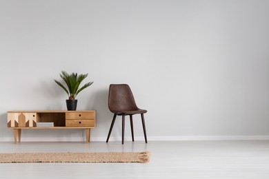 natural linen rug on the floor of spacious bright living room interior with leather chair and wooden cabinet with plant in black pot, real photo with copy space on empty grey wall
