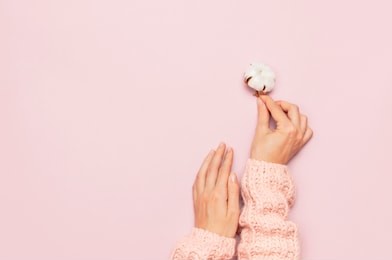 female hands in pink knitted sweater hold a delicate cotton flower on pink pastel table top view. flat lay. copy space. cotton flowers. lifestyle gentle background