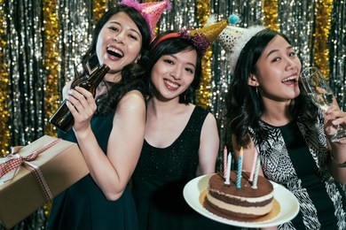 happy friends birthday party at night club. young ladies holding gift and cake having fun in pub. girls with hats enjoy alcohols celebrating event.
