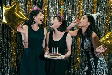 happy friends celebrating birthday with cake in night club. young ladies wearing hats having fun with blower and glod star balloons. beautiful cheerful girl friends laughing together at pub.