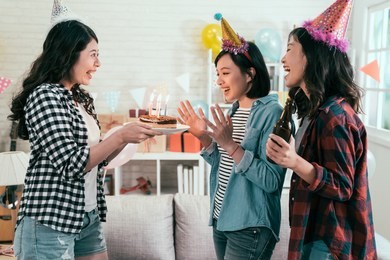 girl best friends birthday party at home singing songs and clapping hands. young ladies giving surprise in the house. women with hats celebrating with chocolate cake and candles.