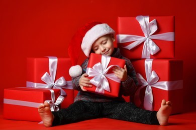 smiling funny child boy in santa red hat fell asleep with christmas gifts in hand in hand on red background. christmas concept.