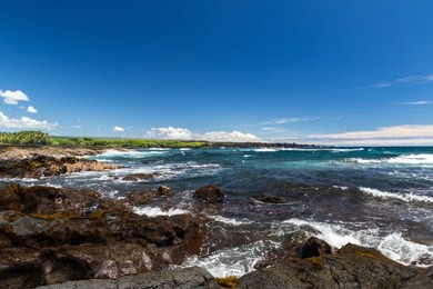 ocean view at black sand beach on hawaii's big island. blue pacific ocean with waves, rough volcanic rock and brown vegetation on shore. coastline with green plants in the distance. 
