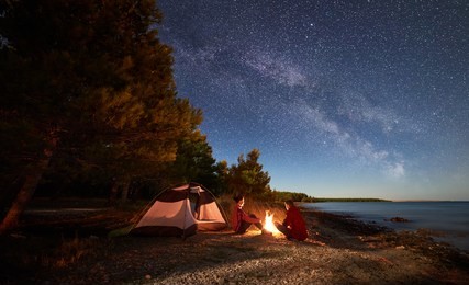 night camping on shore. man and woman hikers having a rest in front of tent at campfire under evening sky full of stars and milky way on blue water and forest background. outdoor lifestyle concept