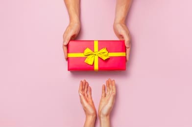 top view of male and female hands holding red gift box with golden ribbon on pink background flat lay. present for birthday, valentine day, christmas, new year. congratulations background copy space.