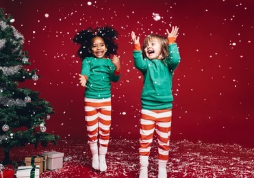 little girls in christmas costume having fun and jumping beside a decorated christmas tree. two kids playing with falling artificial snow flakes near a christmas tree.