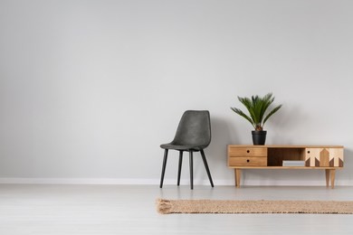 natural linen rug on the floor of spacious bright living room interior with black leather chair and wooden cabinet with plant in black pot, real photo with copy space on empty grey wall