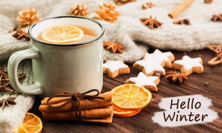 winter theme. cup of hot tea with spices, orange, cinnamon, anise, cookies in a shape of star, fir cones, pepper and gray scarf on wooden background with text hello winter.