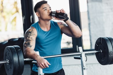muscular asian sportsman drinking water and training with barbell at gym