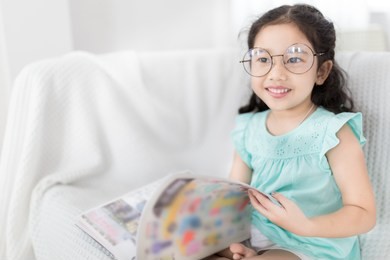 pretty asian children wear glasses and reading a book, education activity, she relax on white sofa